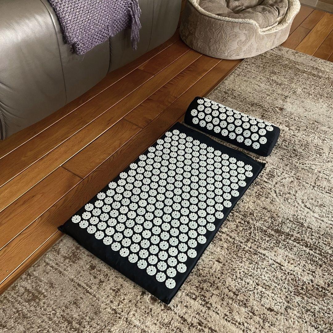 A black acupressure mat set up on a carpet on the floor beside a couch and dog bed, ready for use.